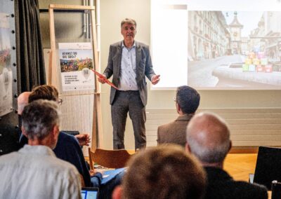 Alec von Graffenried vor Leinwand. Projiziertes Bild zeigt Zytglogge im Hintergrund und kleine SDG-Würfel auf Brunnenrand im Vordergrund.