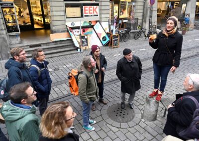 Eva Schmassmann steht auf Sockel und spricht zu Gruppe. Im Hintergrund sieht man Eingang zu Ässbar und Gmüesgarte.