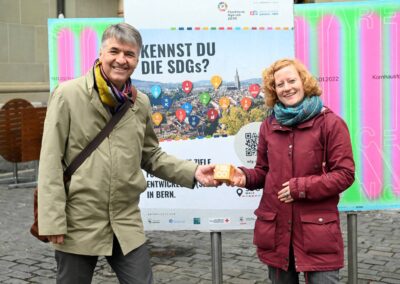 Alec von Graffenried und Karin Spori halten einen kleinen Papierwürfel mit SDG 12-Icon. Im Hintergrund Plakat des SDG Walk.