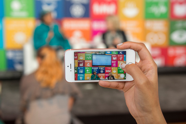 Eine Hand hält ein Handy. Auf dem Handybildschirm sieht man 2 Personen auf einer Bühne. Im Hintergrund die SDGs.